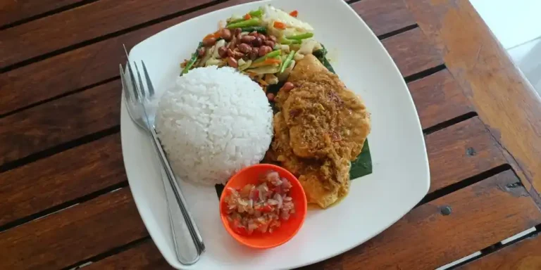 Balinese Ayam Betutu served with white rice, stir-fried vegetables, peanuts and sambal on a white plate.