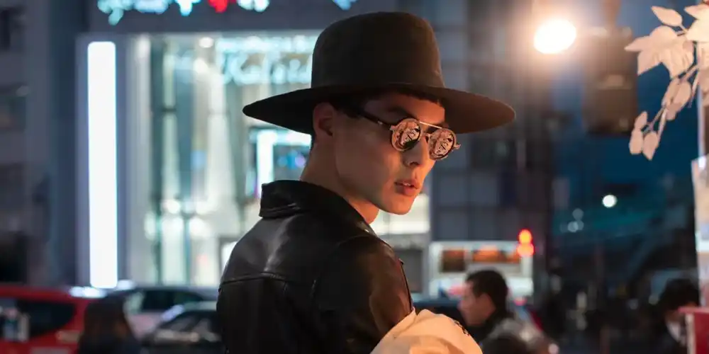 Young man wearing a wide-brimmed hat, round gold glasses and a black leather jacket while carrying a Chinese character printed garment on a city street in China.
