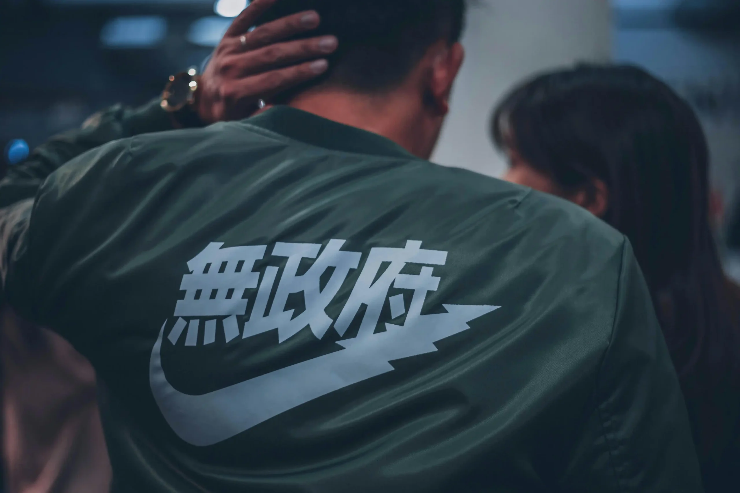 Pria mengenakan jaket gelap dengan aksara China berukuran besar dan logo Nike di bagian belakang.
