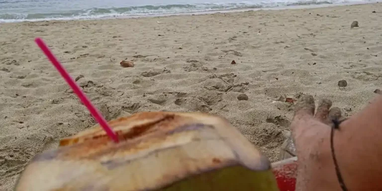 Coconut drink with a pink straw on the sand at Mawun Beach, with green hills and calm sea in the background.
