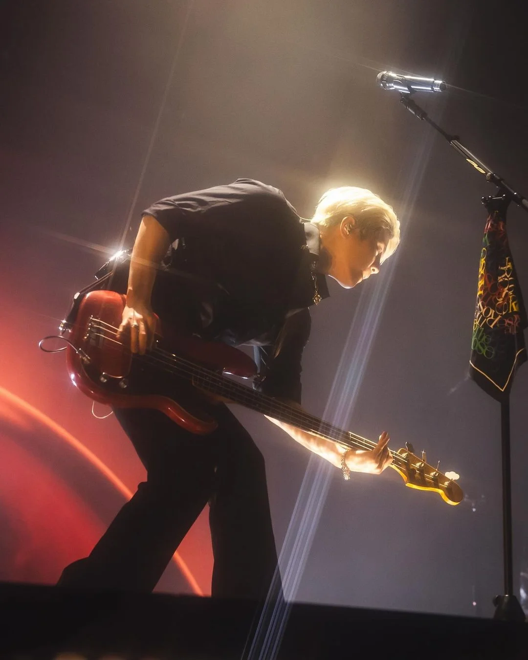A member of The Rose playing bass guitar on stage while leaning forward beneath warm concert lights.