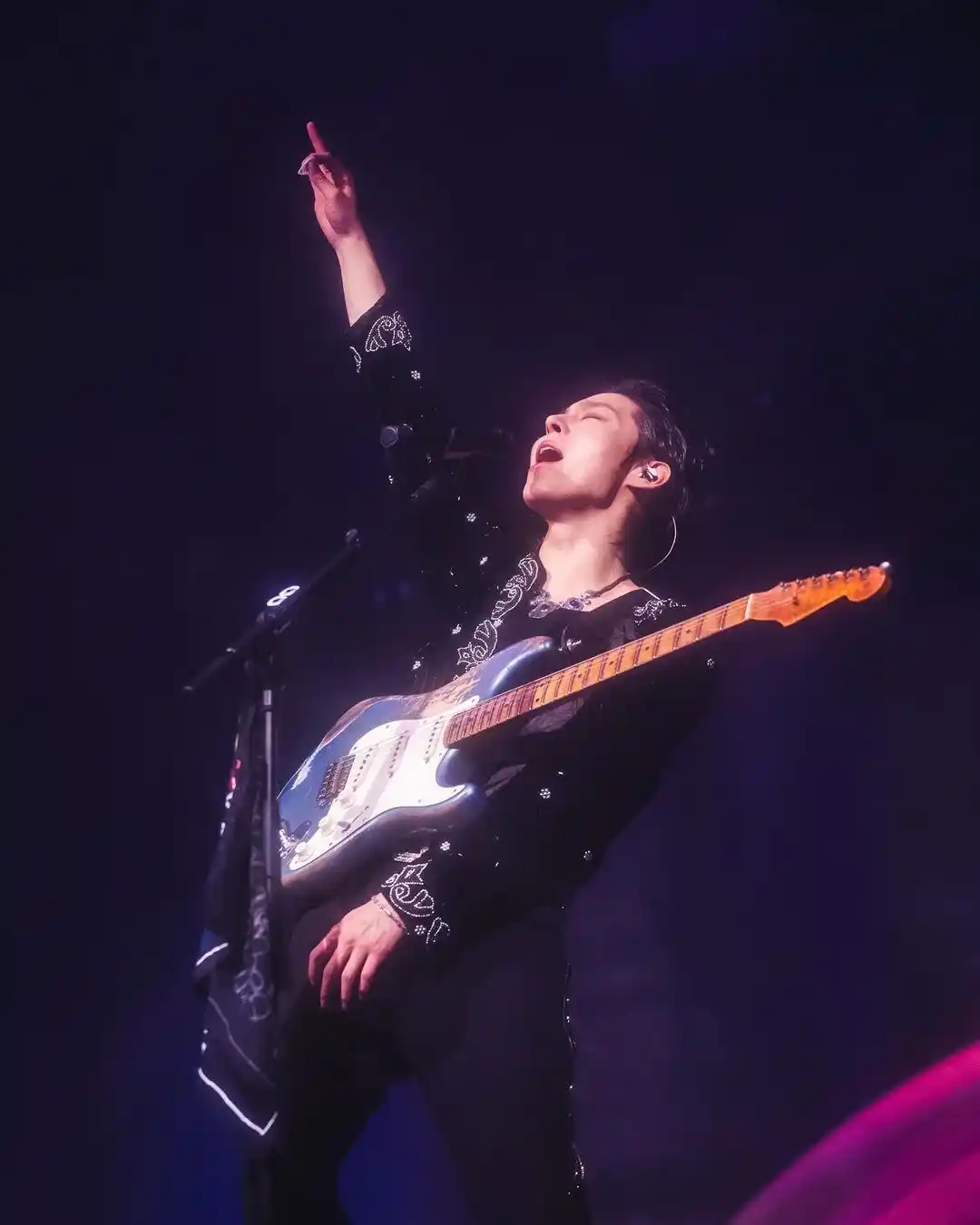 A member of The Rose singing and playing electric guitar on stage with one hand raised under purple concert lighting.