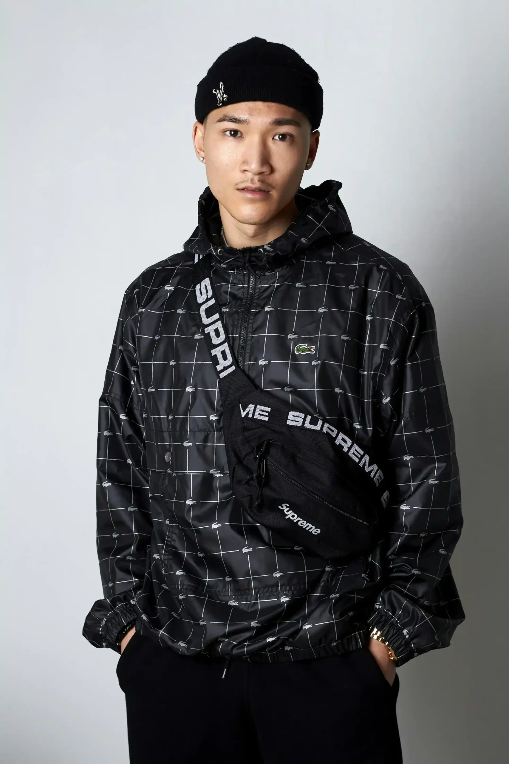 Pria muda mengenakan jaket hitam bermotif dengan hoodie, celana hitam, beanie hitam dan tas selempang hitam.