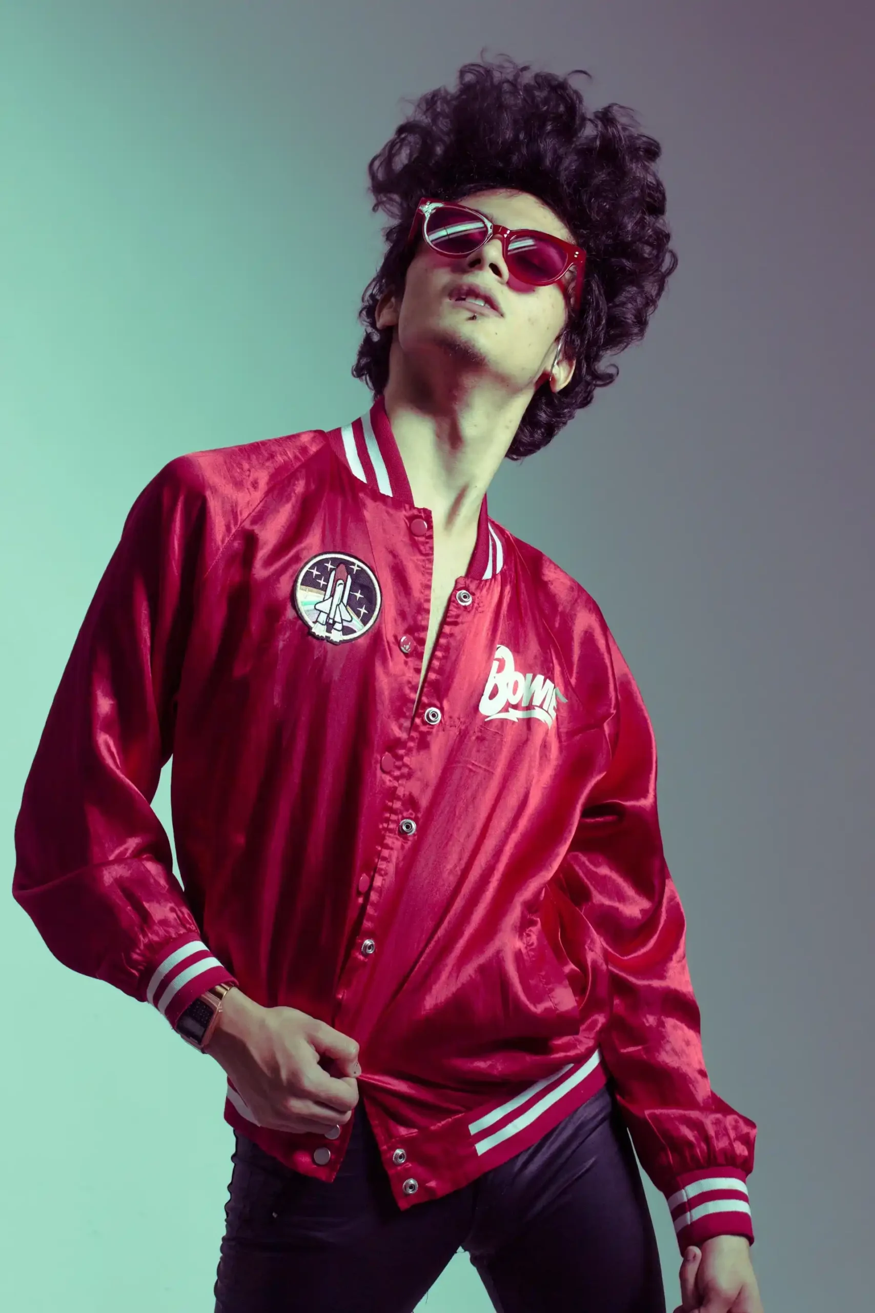 Pria muda mengenakan jaket varsity satin merah, kacamata hitam merah dan celana gelap dengan gaya percaya diri.