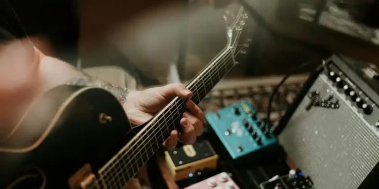 Close-up of a guitarist playing an electric guitar beside guitar pedals and an amplifier in a music studio.