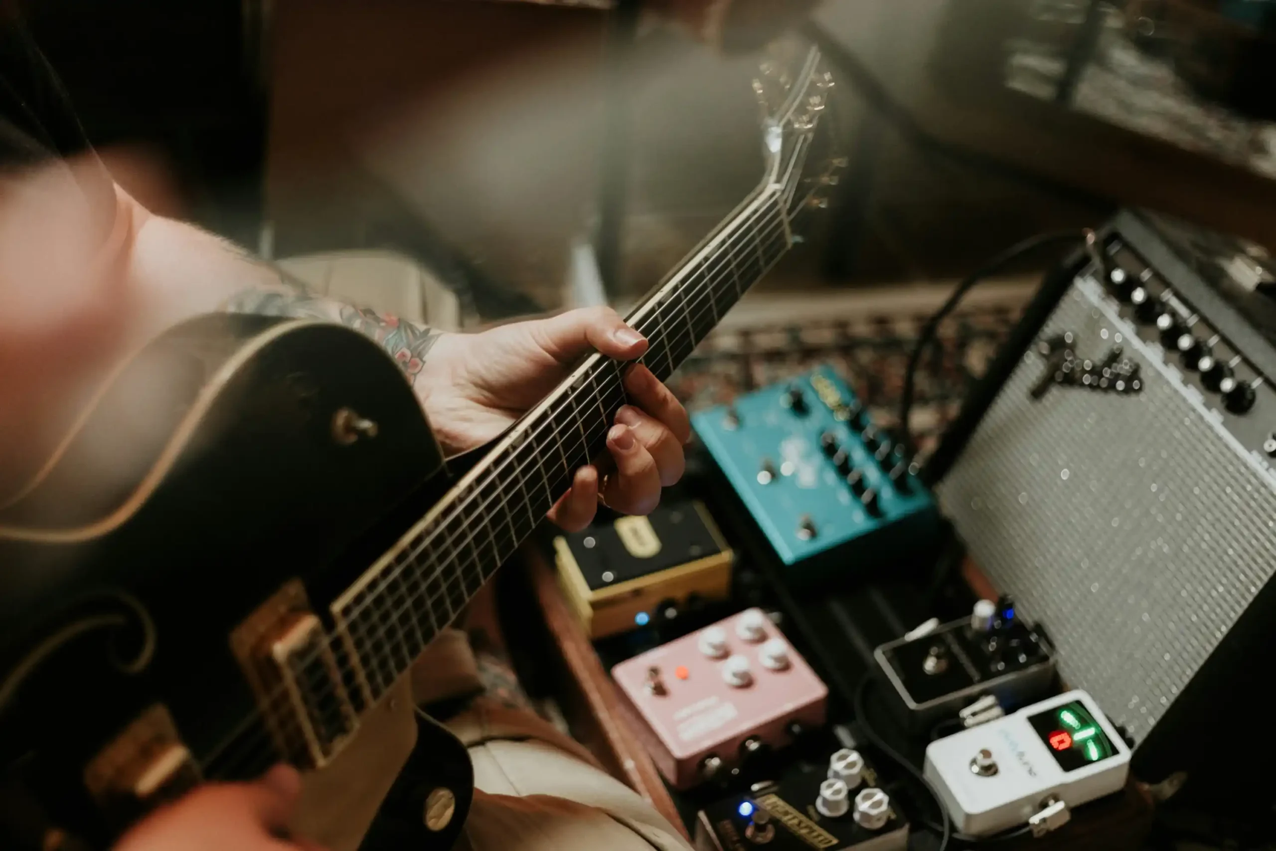 Close-up of a guitarist playing an electric guitar beside guitar pedals and an amplifier in a music studio.