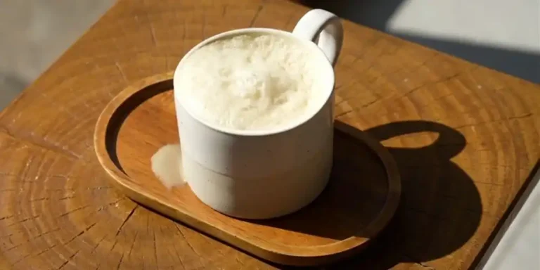 A white ceramic cup of frothy butter tea sits on a wooden tray atop a rustic wooden table, with a small spill beside it in warm sunlight.