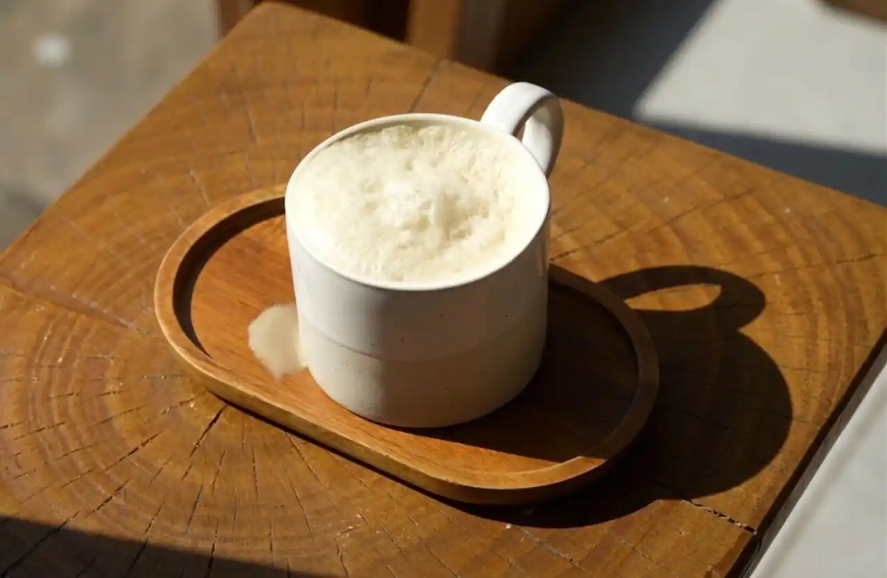 A white ceramic cup of frothy butter tea sits on a wooden tray atop a rustic wooden table, with a small spill beside it in warm sunlight.
