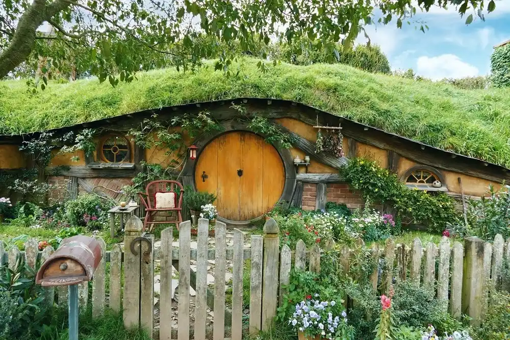 Traditional hobbit house at the Hobbiton film set in New Zealand with a round wooden door, grass-covered roof, garden flowers and white picket fence.