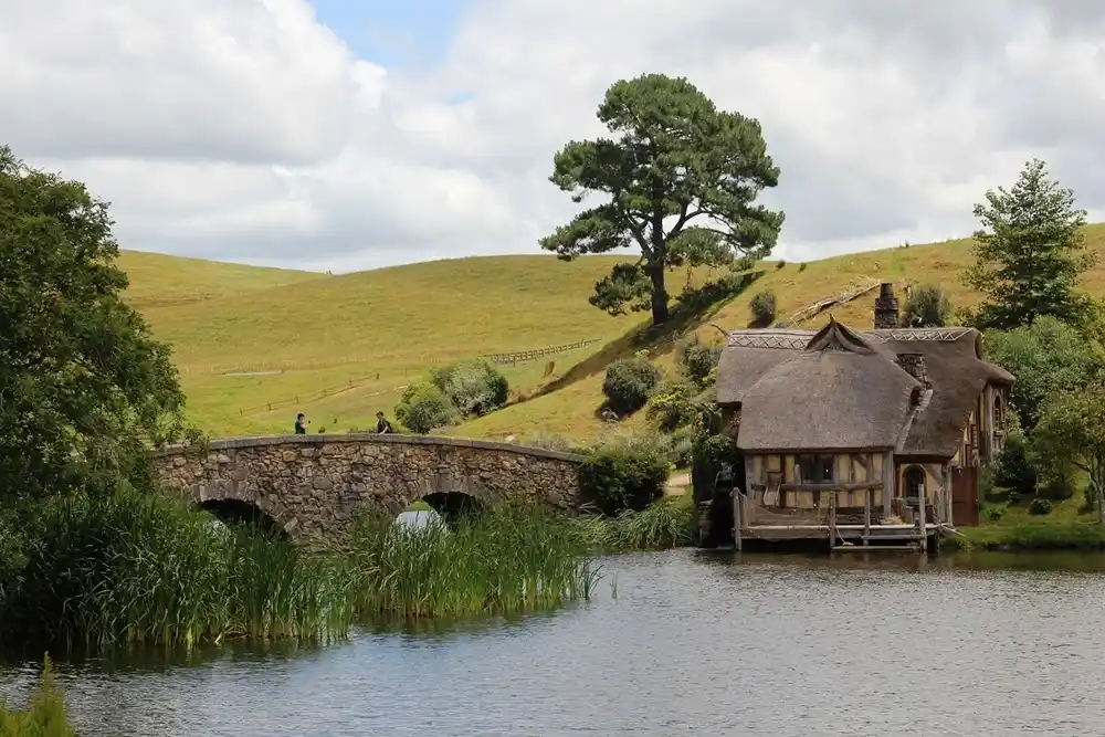 Hobbiton film set in Matamata, New Zealand, featuring a stone bridge, lakeside hobbit house and green rolling hills beneath cloudy skies.