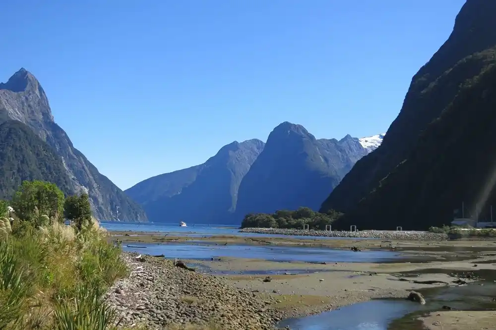 Milford Sound in Fiordland National Park, New Zealand, with dramatic mountain cliffs, calm water and rocky shoreline beneath a clear blue sky.