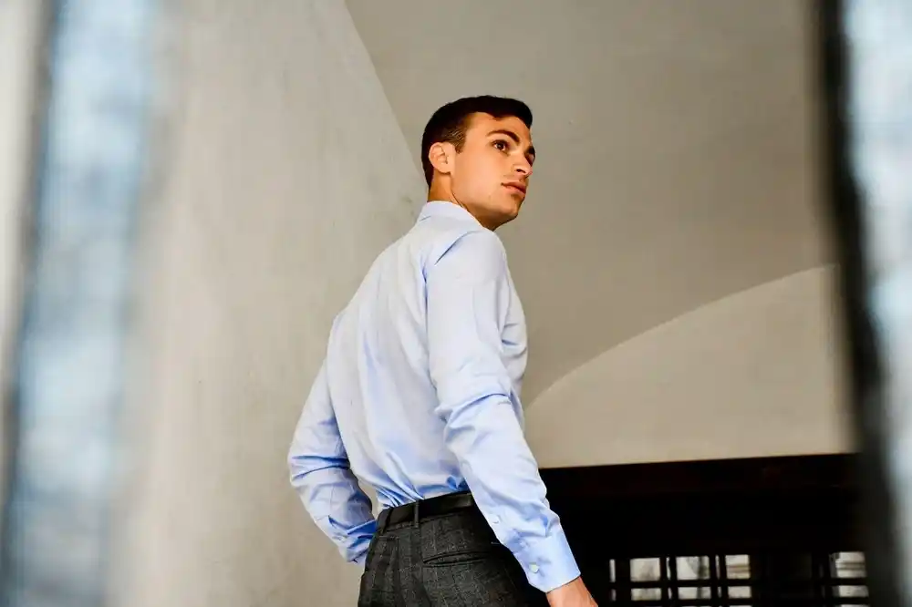 Man wearing a light blue long-sleeved shirt tucked into grey trousers, standing on a staircase.