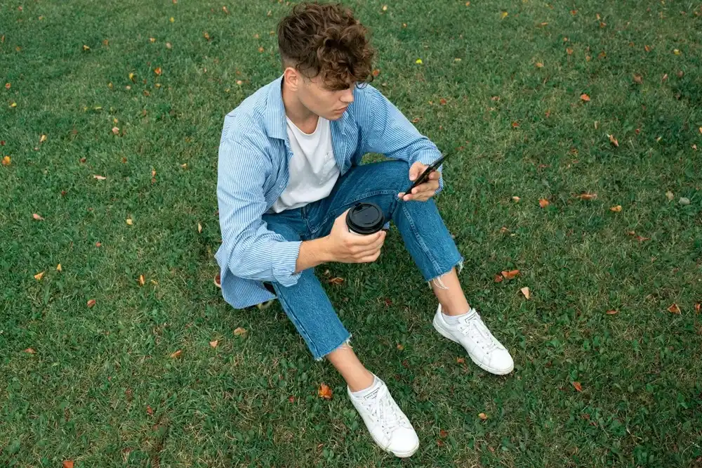 Man wearing a blue striped shirt over a white T-shirt with blue jeans and white trainers, sitting on the grass while holding a takeaway coffee and a smartphone.