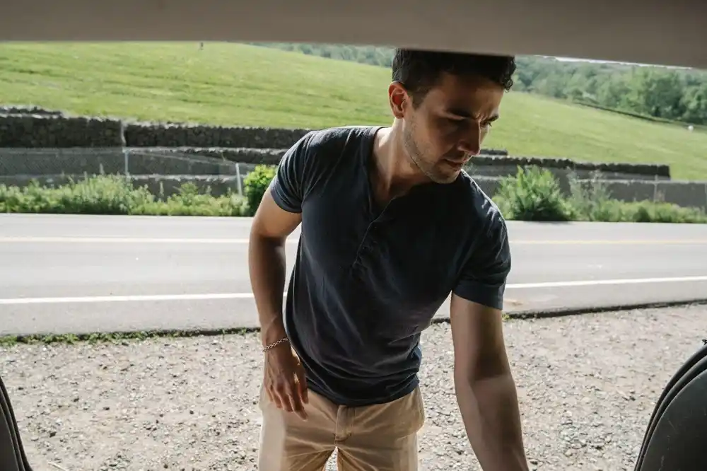 Man wearing a dark Henley shirt with beige chinos, reaching into the boot of a car beside a country road.