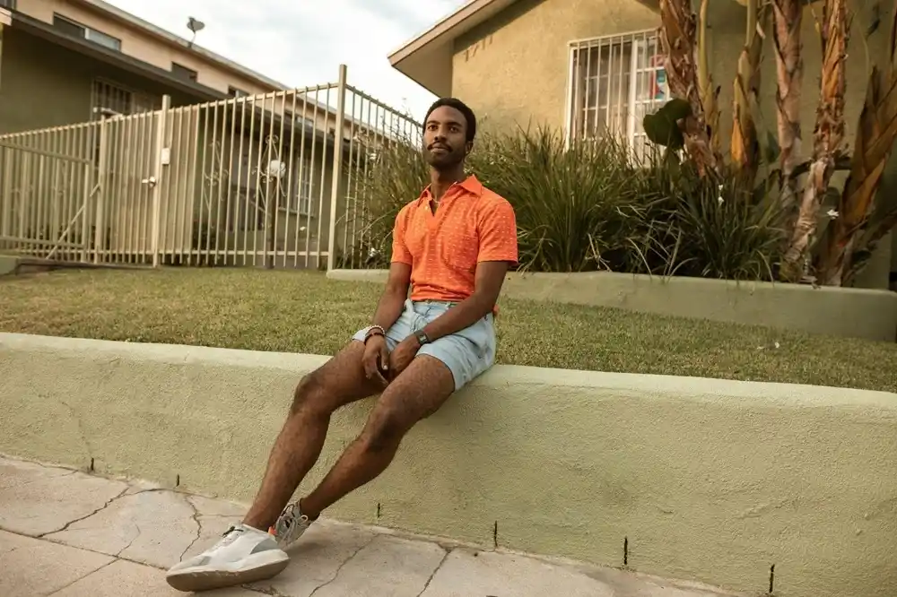 Man wearing an orange polo shirt with light blue denim shorts and white trainers, sitting on a low wall outside a house.
