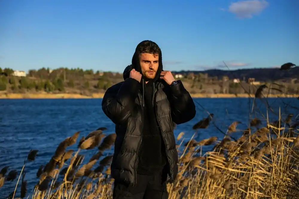 Man wearing a black hooded puffer jacket, standing beside a lake on a cold winter day.