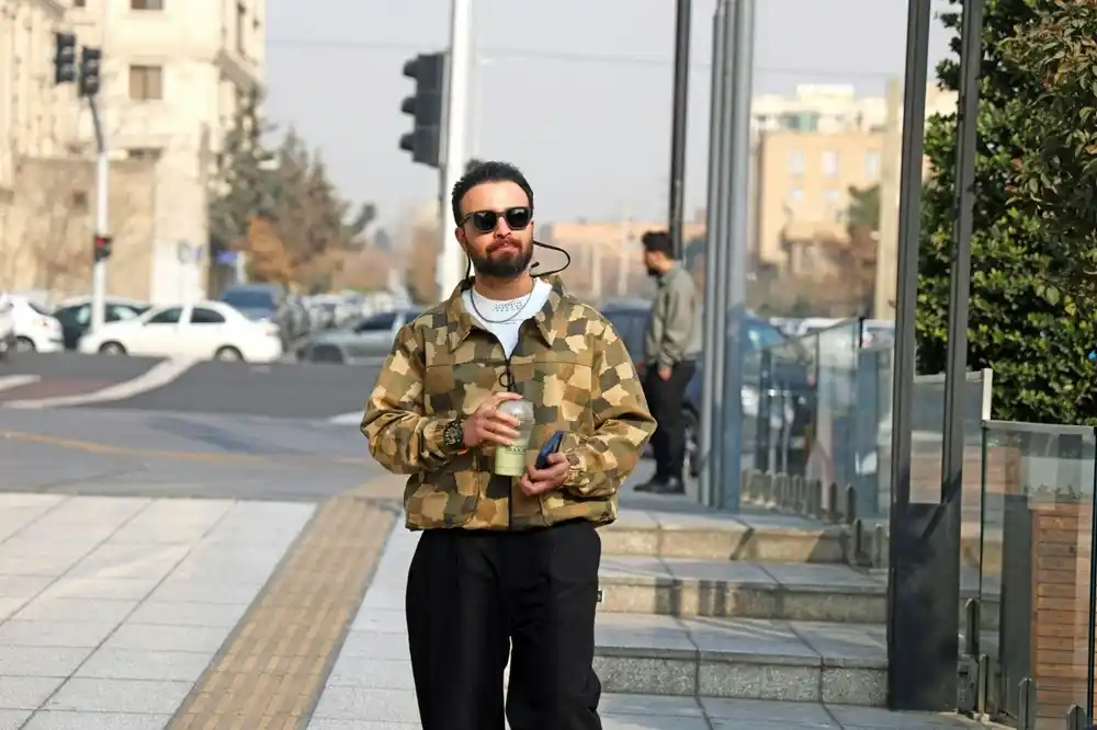 Man wearing a camouflage-print jacket over a white T-shirt with black trousers, walking along a city street while holding a takeaway drink.