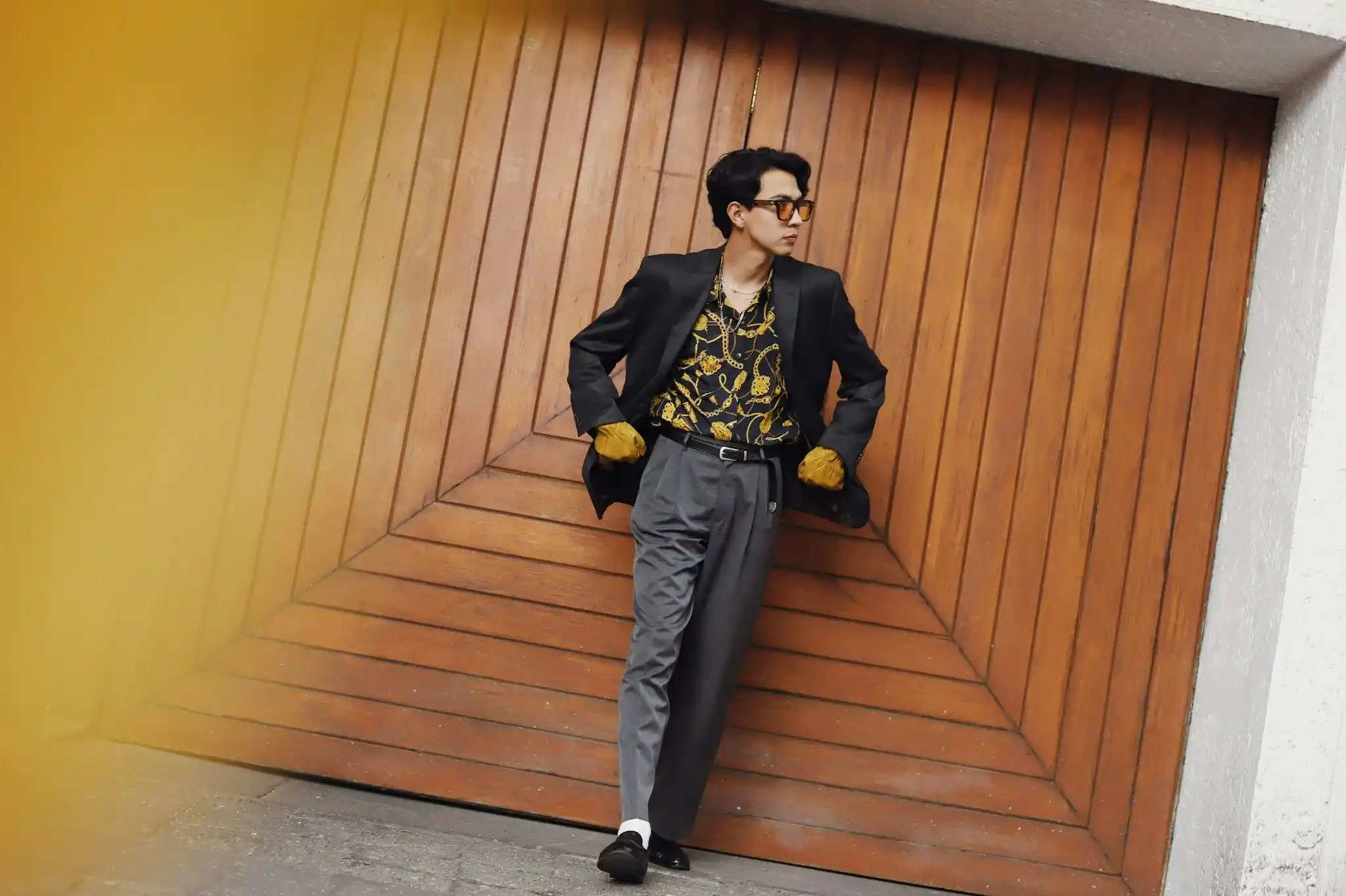 Man wearing a black blazer over a patterned shirt with grey trousers and loafers, leaning against a large wooden door.