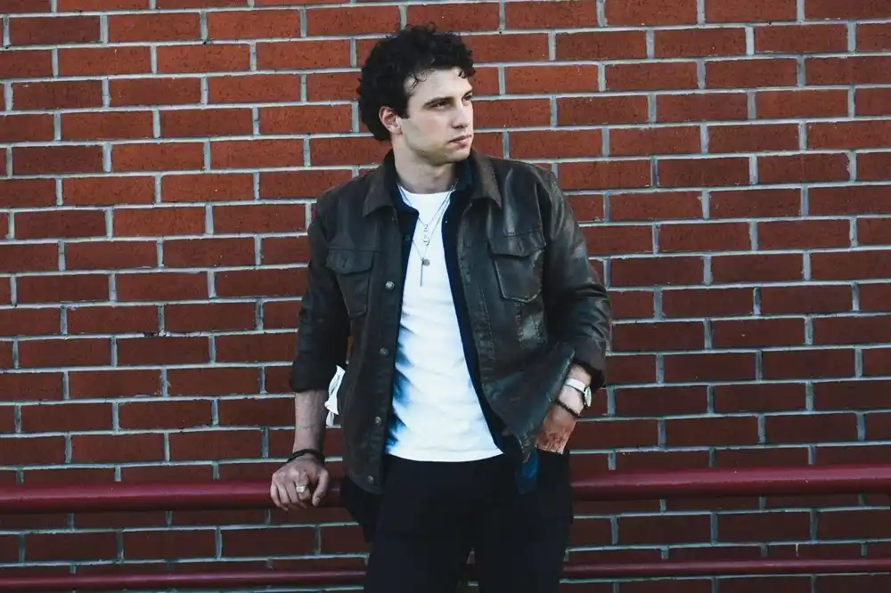 Man wearing a dark leather jacket over a white T-shirt with dark trousers, standing against a red brick wall.
