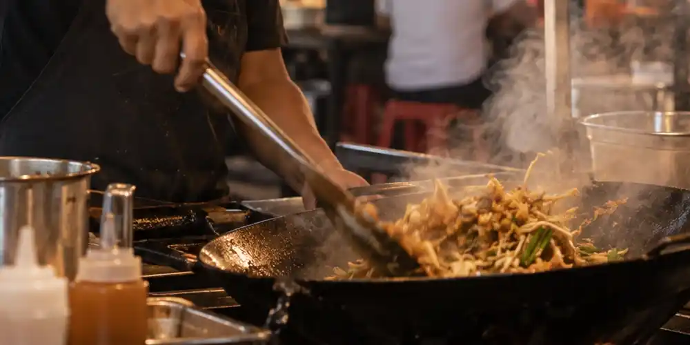 Street food vendor cooking noodles in a hot wok at a busy Southeast Asian night market.