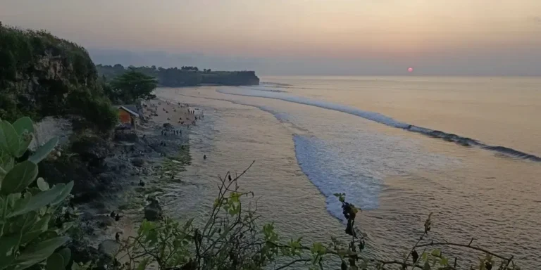 Sunset over Balangan Beach in Bali, with gentle waves rolling towards the shore, rocky cliffs covered in greenery and people gathered along the beach.